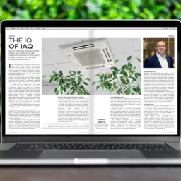 Laptop on a wooden surface displaying a magazine article titled 'The IQ of IAQ' with a photo of a ceiling air conditioning unit and green plants. Background includes green foliage.