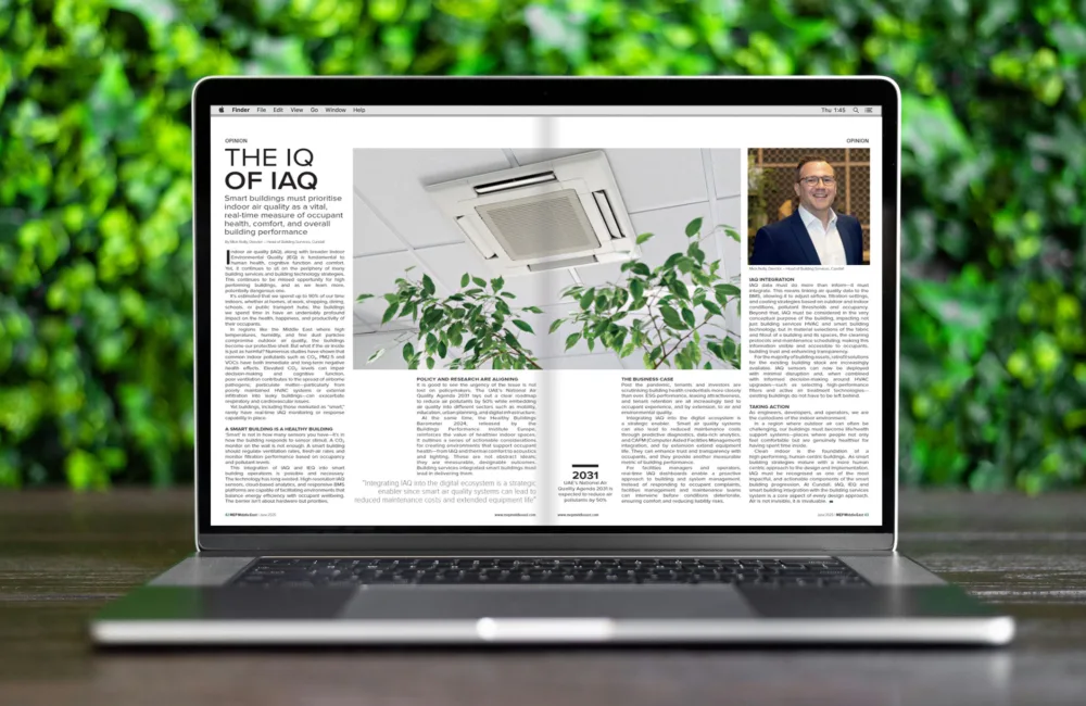 Laptop on a wooden surface displaying a magazine article titled 'The IQ of IAQ' with a photo of a ceiling air conditioning unit and green plants. Background includes green foliage.