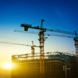 Construction site at sunrise with multiple tower cranes above partially built structures covered in scaffolding, set against a golden sky and deep blue horizon.