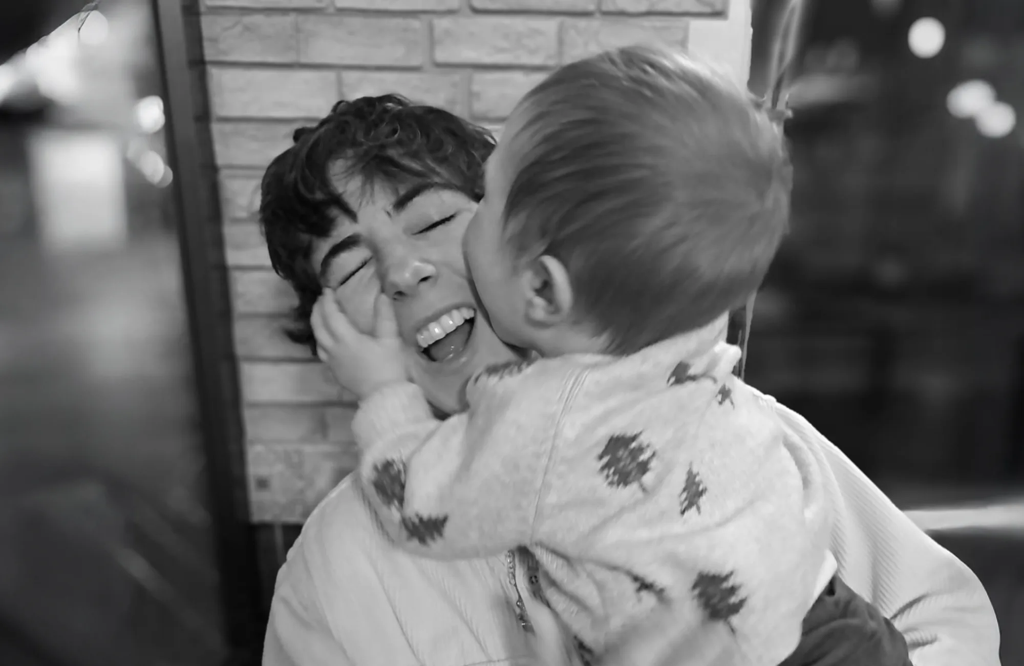 Black and white image of Lucia smiling joyfully with a small child on her lap kissing her cheek.