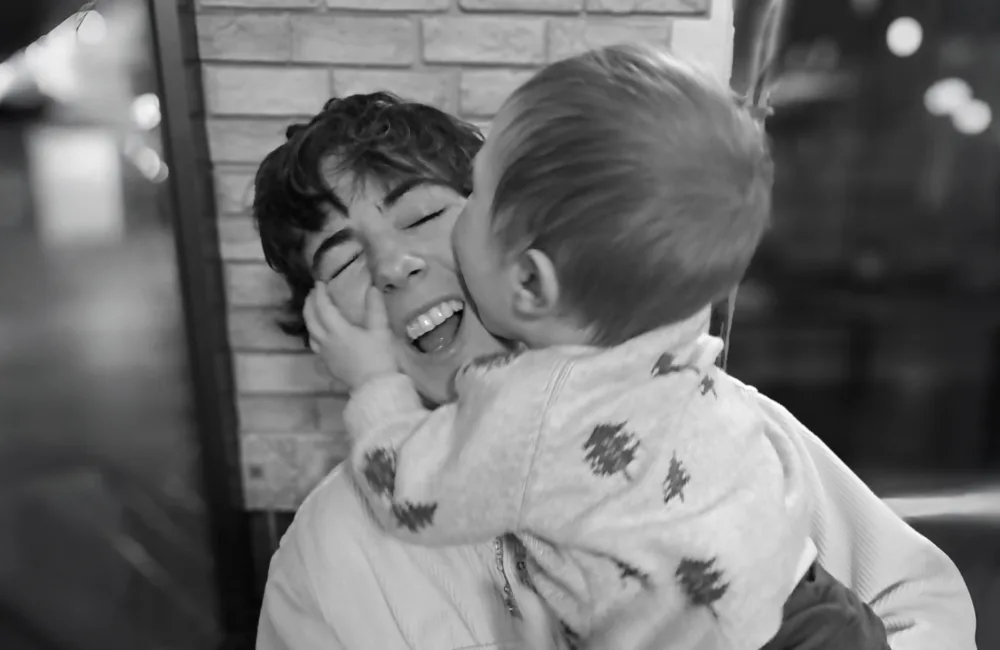 Black and white image of Lucia smiling joyfully with a small child on her lap kissing her cheek.