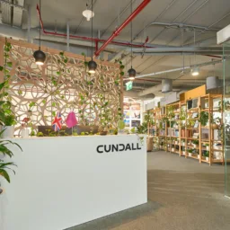 Modern office reception area with a white front desk labelled “CUNDALL”, indoor plants in pots, decorative wood screen with greenery, overhead exposed ceiling pipes, shelving units, glass partition with mounted project images, and carpeted flooring.