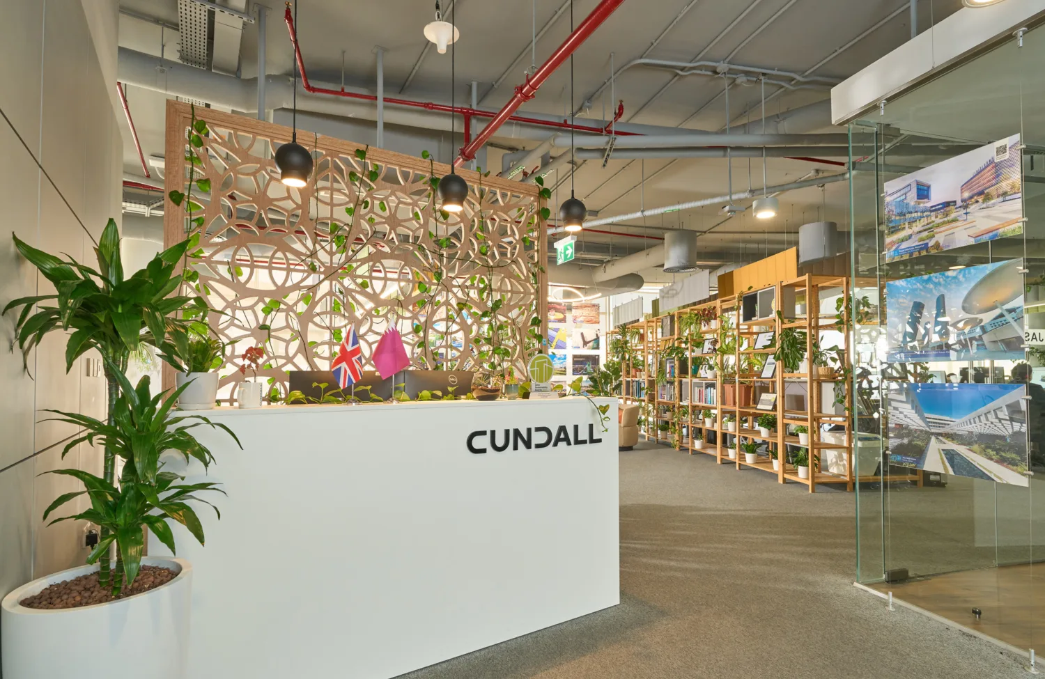Modern office reception area with a white front desk labelled “CUNDALL”, indoor plants in pots, decorative wood screen with greenery, overhead exposed ceiling pipes, shelving units, glass partition with mounted project images, and carpeted flooring.