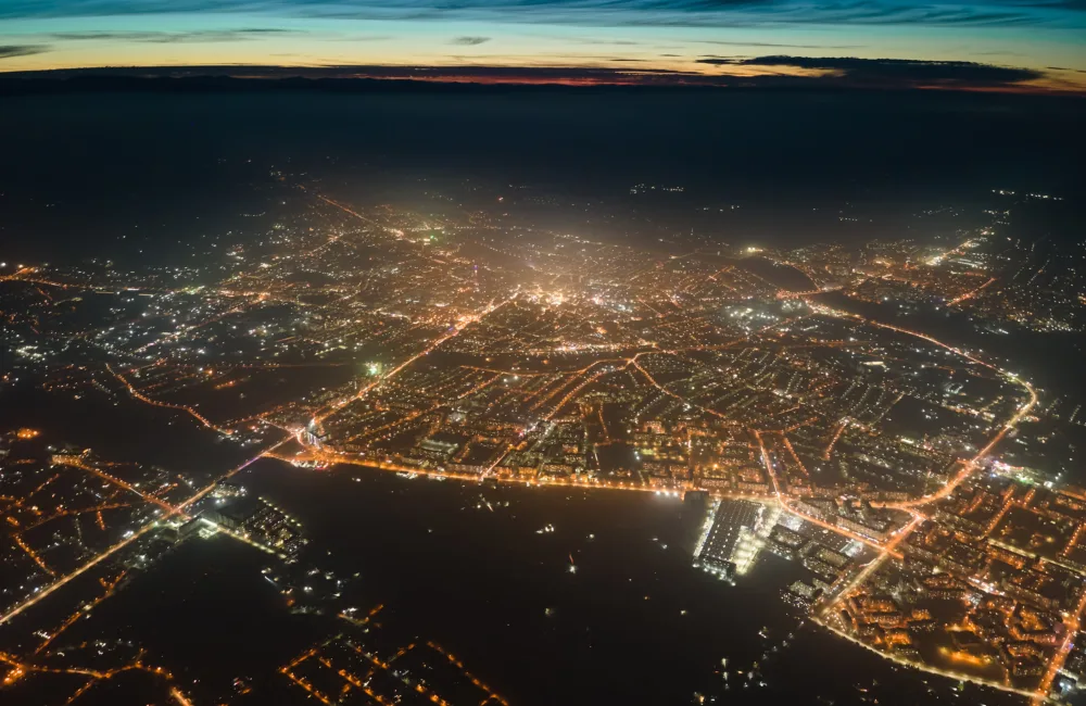 Aerial view of a city at night with illuminated streets forming a grid pattern around dark water areas. Dense clusters of lights mark buildings, with a glowing horizon and layered clouds above.