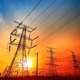 Silhouetted high-voltage power lines and transmission towers against a vivid sunset sky with orange, red, and blue hues; sun partially visible near the horizon.