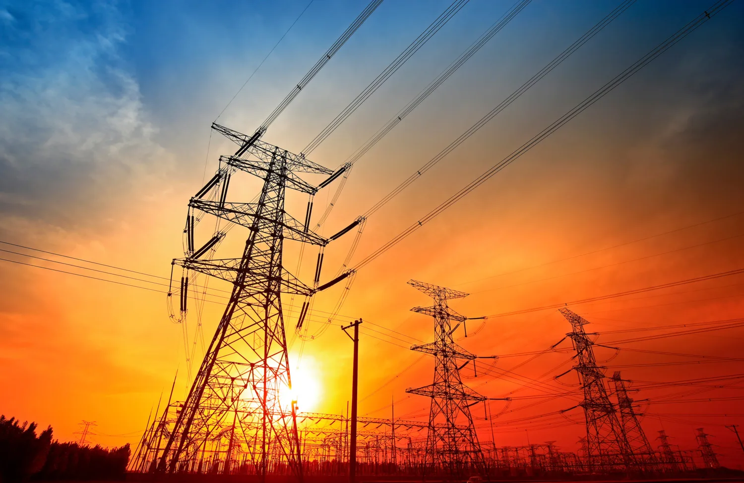 Silhouetted high-voltage power lines and transmission towers against a vivid sunset sky with orange, red, and blue hues; sun partially visible near the horizon.