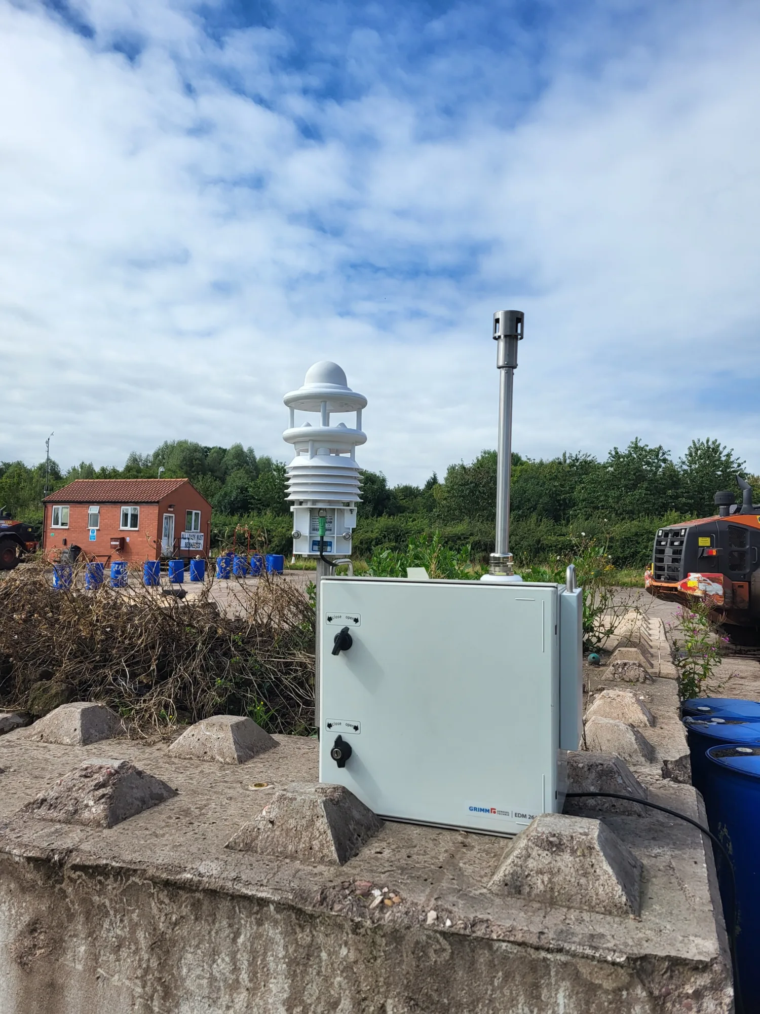 Outdoor scene with a white weather‑monitoring instrument, a tall sensor unit, and a light grey equipment box on a concrete surface. A red brick building, vegetation, machinery, and a cloudy sky appear in the background.
