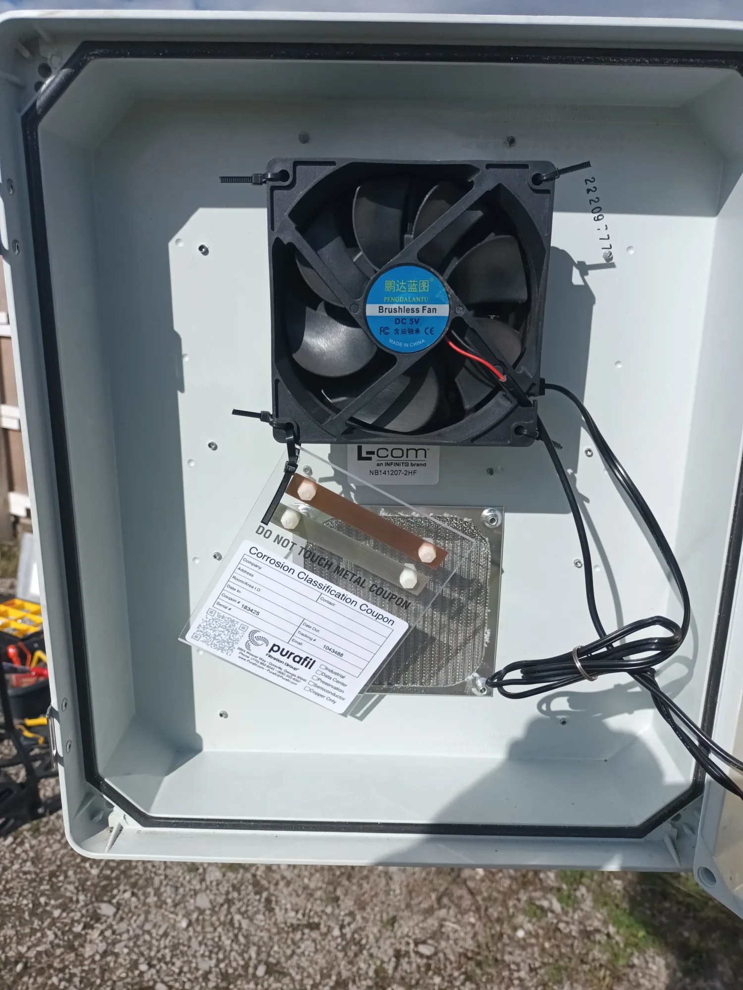 Open equipment cabinet showing an internal cooling fan, wiring, and a mounted metal plate holding a labelled test coupon. The cabinet is outdoors on a gravel surface with tools and structures visible in the background.