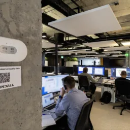 Cundall Edinburgh office with three people working at desks with multiple monitors. A wall-mounted device shows '878 ppm' and '82 dB'. A sign below reads 'Check the indoor air quality here' with a QR code and 'CUNDALL'.