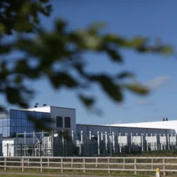 Large modern building with white walls and reflective glass panels, surrounded by metal fencing and greenery. A wooden fence runs along the foreground, and tree branches partially frame the view under a clear blue sky.