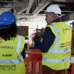 Two people wearing high-visibility vests and hard hats stand at a construction site behind orange safety barriers. The vest on the left reads “Cundall Graduate Protecting People,” and the vest on the right reads “Overbury Senior Technic Services Manager Protecting People.” Visible scaffolding and equipment are in the background.