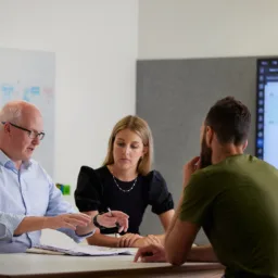 Three people seated around a table in a meeting room with documents on the table. A whiteboard with diagrams is on the left and a large screen displaying a technical schematic is on the right.