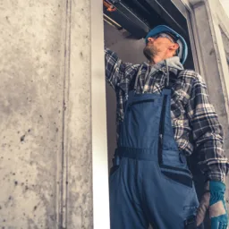 A worker wearing blue overalls, gloves, and a hard hat stands in a concrete-walled area, reaching up to inspect or repair equipment inside an open doorway or shaft.