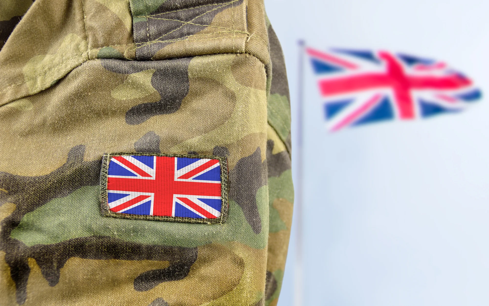Close-up of a camouflage military uniform with a stitched Union Jack patch on the sleeve. In the blurred background, a larger Union Jack flag is visible against a light gray backdrop.