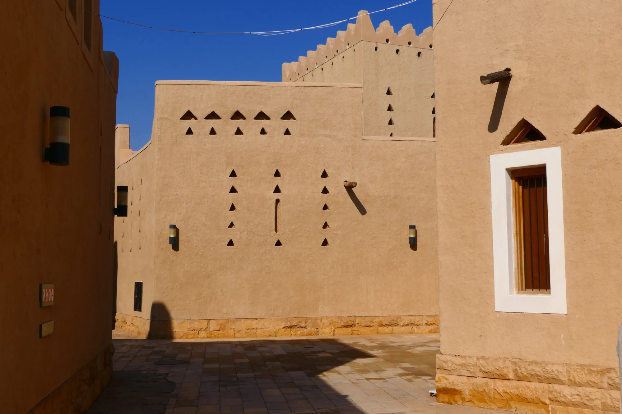 a {     text-decoration: none;     color: #464feb; } tr th, tr td {     border: 1px solid #e6e6e6; } tr th {     background-color: #f5f5f5; }   Sunlit alley between sand-colored buildings with geometric cut‑out patterns, wooden window frames, wall-mounted lights, and a paved stone walkway under a clear blue sky.