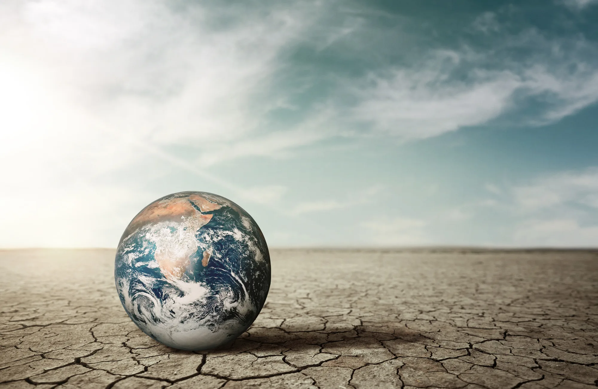 A globe of Earth rests on cracked, dry ground beneath a partly cloudy sky. Sunlight enters from the left, casting shadows on the terrain. No people or text are visible.