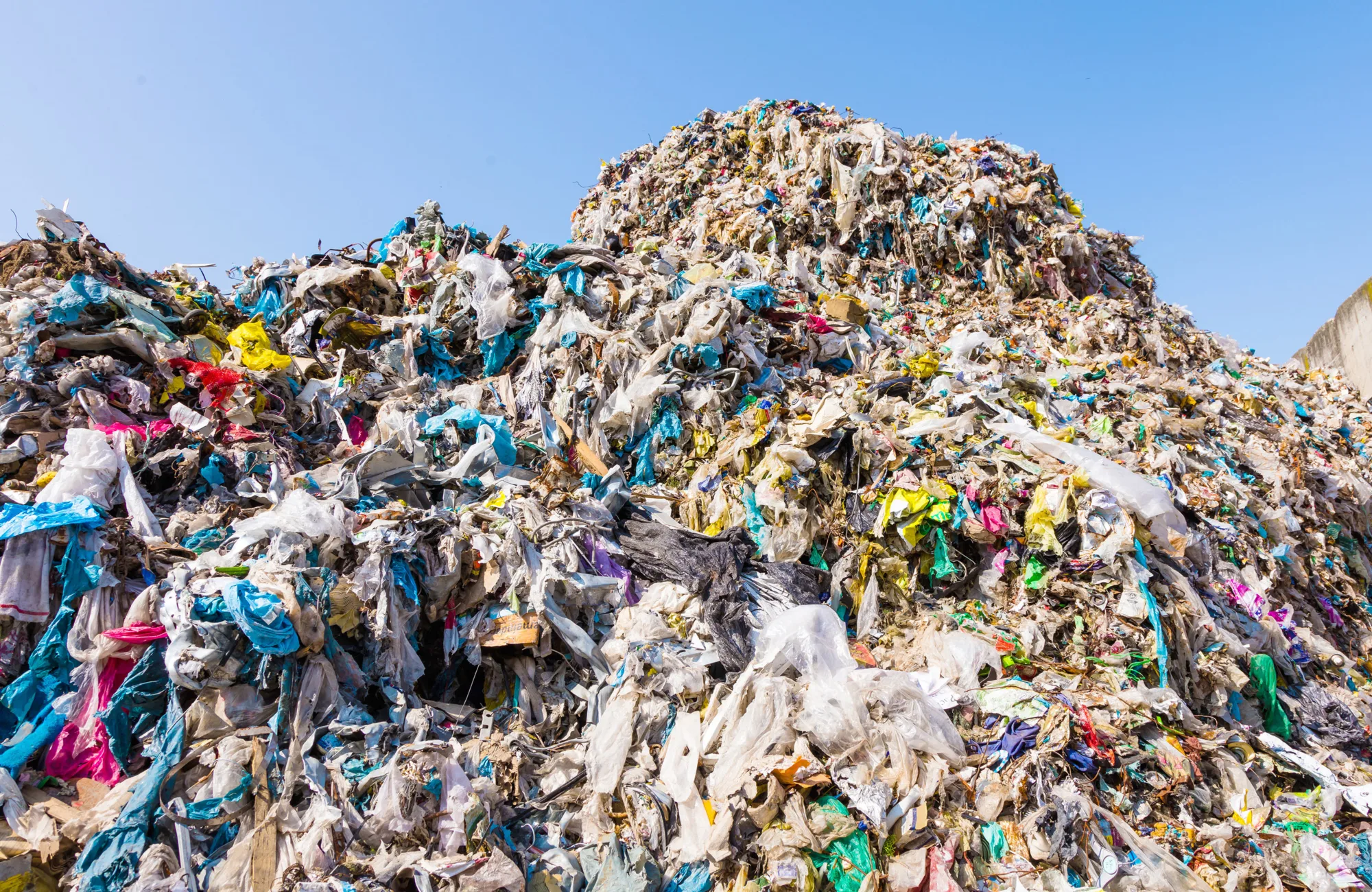A large, densely packed pile of mixed waste, mainly plastic bags in various colors, sits outdoors beneath a clear blue sky. No people or text are visible in the image.