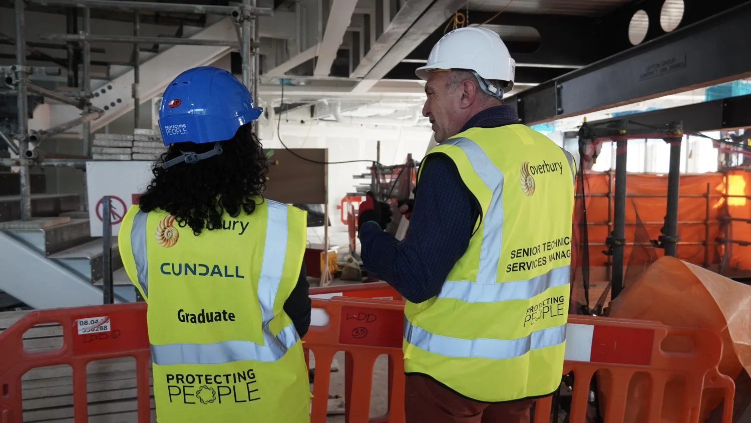 Two people wearing high-visibility vests and hard hats stand at a construction site behind orange safety barriers. The vest on the left reads “Cundall Graduate Protecting People,” and the vest on the right reads “Overbury Senior Technic Services Manager Protecting People.” Visible scaffolding and equipment are in the background.