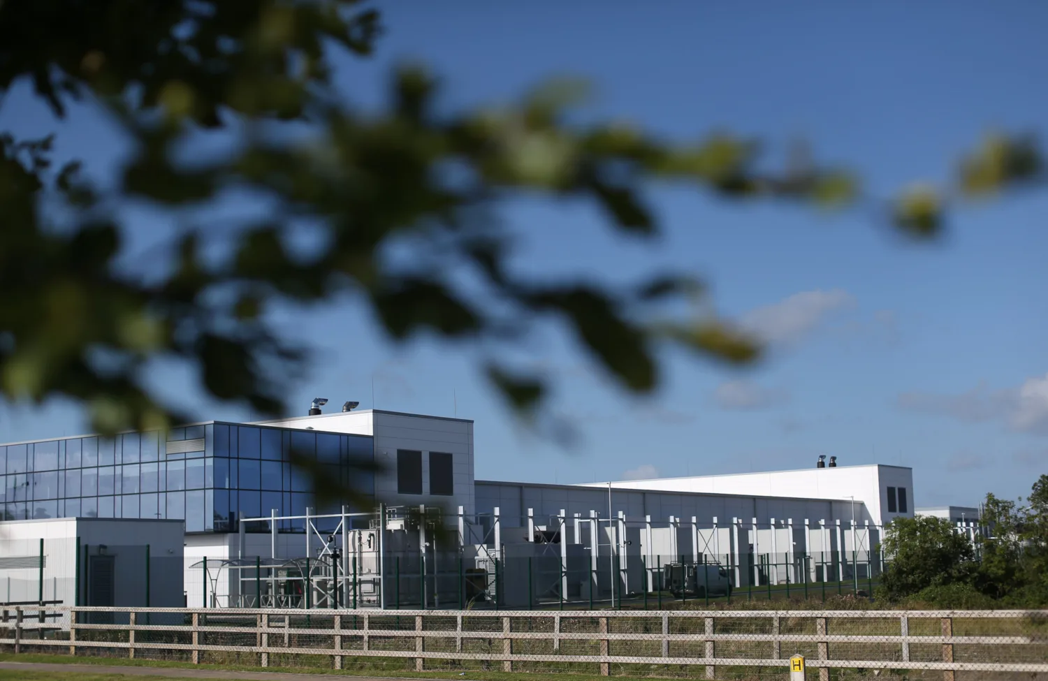 Large modern building with white walls and reflective glass panels, surrounded by metal fencing and greenery. A wooden fence runs along the foreground, and tree branches partially frame the view under a clear blue sky.