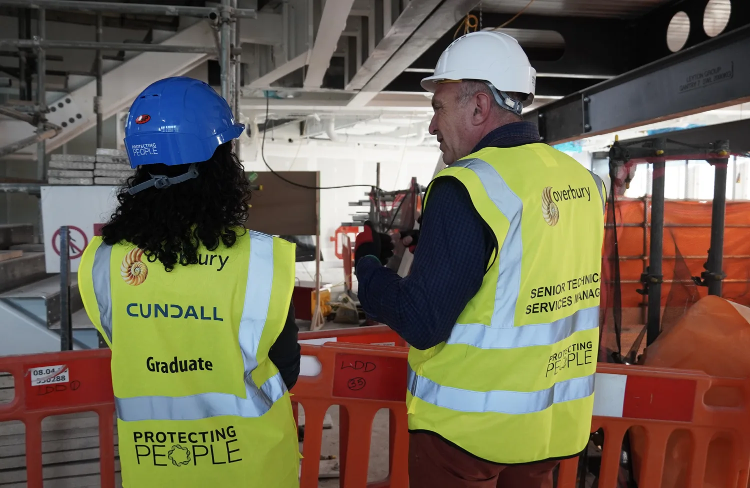 Two people wearing high-visibility vests and hard hats stand at a construction site behind orange safety barriers. The vest on the left reads “Cundall Graduate Protecting People,” and the vest on the right reads “Overbury Senior Technic Services Manager Protecting People.” Visible scaffolding and equipment are in the background.