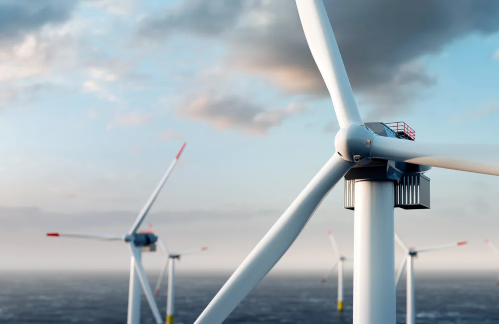 A close-up view of a wind turbine with several other wind turbines in the background, situated in an offshore wind farm. The sky is partly cloudy, and the sea is visible below the turbines.