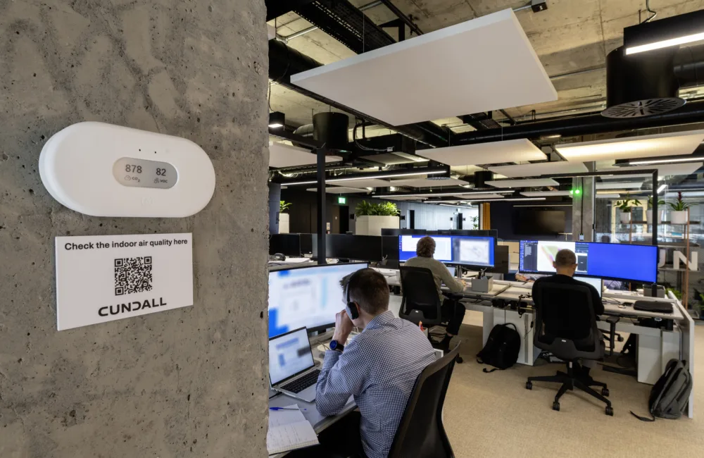 Cundall Edinburgh office with  three people working at desks with multiple monitors. A wall-mounted device shows '878 ppm' and '82 dB'. A sign below reads 'Check the indoor air quality here' with a QR code and 'CUNDALL'.