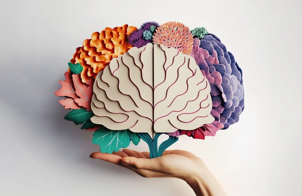 A hand holds a stylized paper-cutout brain with detailed folds and veins, surrounded by textured paper flowers in orange, pink, purple, and green, set against a plain white background.