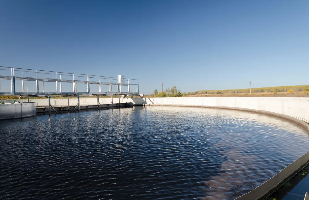 A circular water treatment basin filled with clear blue water, bordered by a concrete wall. A metal walkway with railings runs along the left side. Trees and open land are visible in the background.