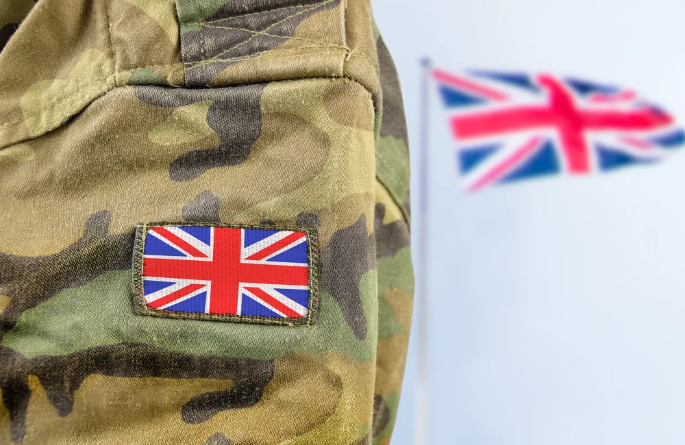 Close-up of a camouflage military uniform with a stitched Union Jack patch on the sleeve. In the blurred background, a larger Union Jack flag is visible against a light gray backdrop.