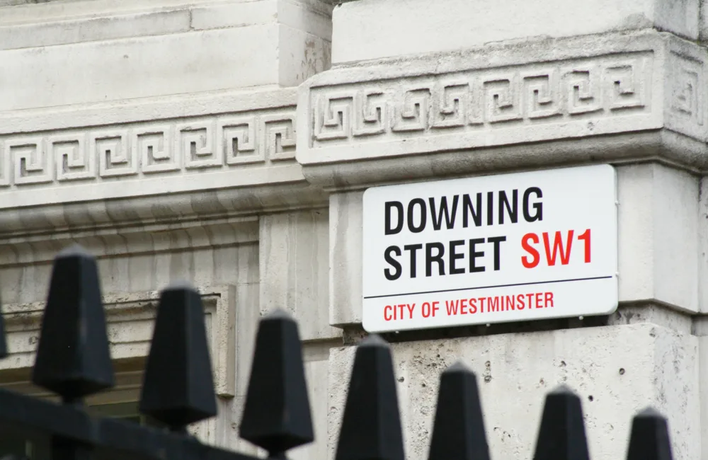 Close-up of a white street sign mounted on a stone building facade with decorative geometric carvings. The sign reads “DOWNING STREET SW1” in black and red letters, with “CITY OF WESTMINSTER” in smaller red text below. Black metal railings are visible in the foreground.