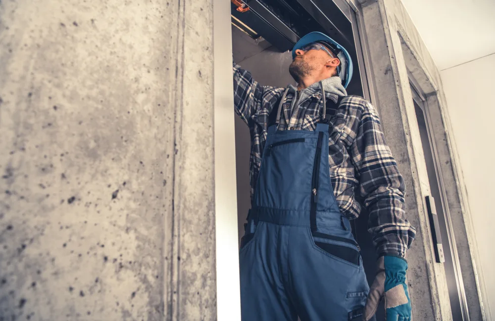 A worker wearing blue overalls, gloves, and a hard hat stands in a concrete-walled area, reaching up to inspect or repair equipment inside an open doorway or shaft.