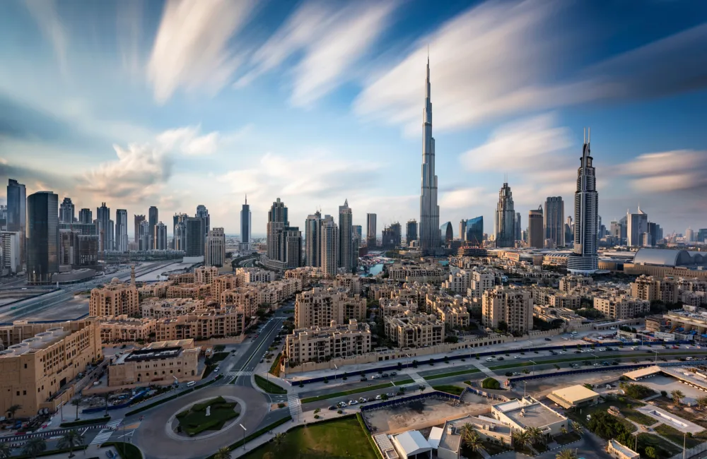 A panoramic daytime view of a modern city skyline with numerous high-rise towers and dense low-rise buildings in the foreground. A tall, slender skyscraper stands prominently at the center under a sky with long, streaked clouds.