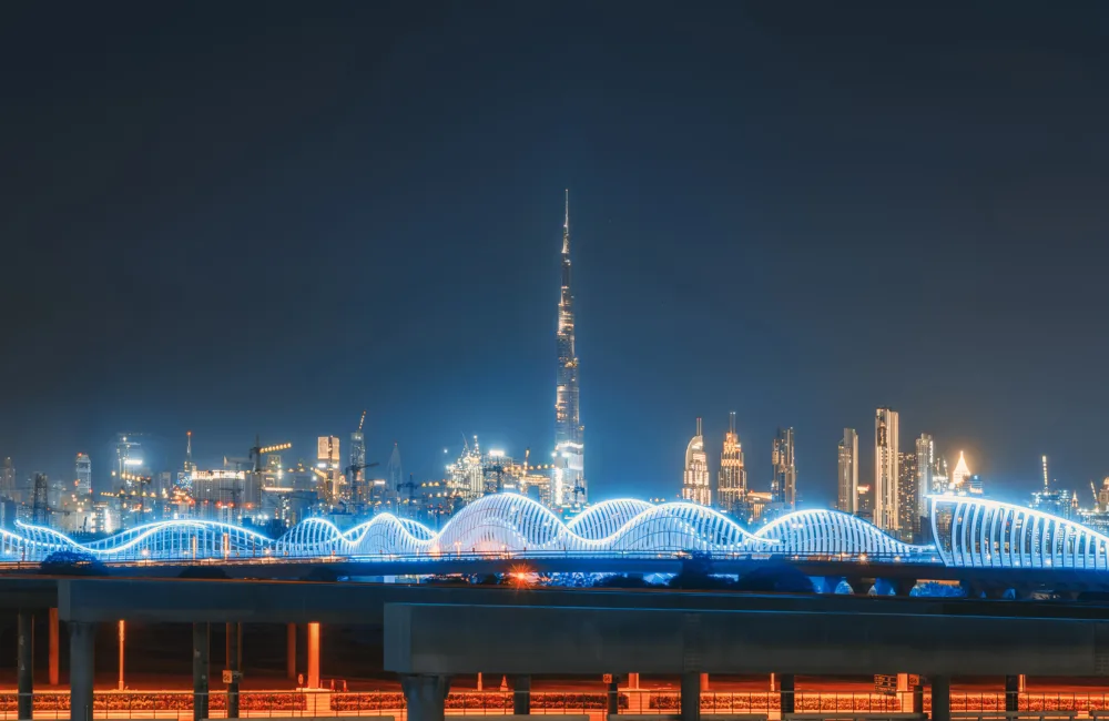 A nighttime cityscape featuring a brightly illuminated wavy bridge in the foreground and a skyline of tall modern towers in the background, all lit against a dark sky.