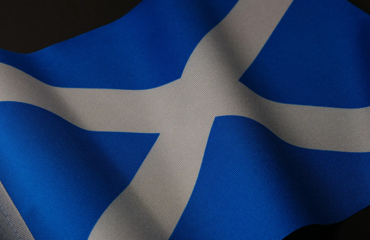 Close-up of the Scottish flag showing a white diagonal cross on deep blue fabric, with visible woven texture and soft folds creating light and shadow across the cloth.
