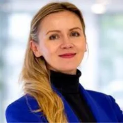 Head-and-shoulders portrait of a person wearing a bright blue blazer over a black high‑neck top, with long light‑coloured hair pulled back, set against a softly blurred indoor office background with natural light.