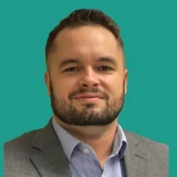 Head-and-shoulders studio portrait of a person wearing a grey suit jacket over a light blue collared shirt, positioned centrally against a solid teal background.