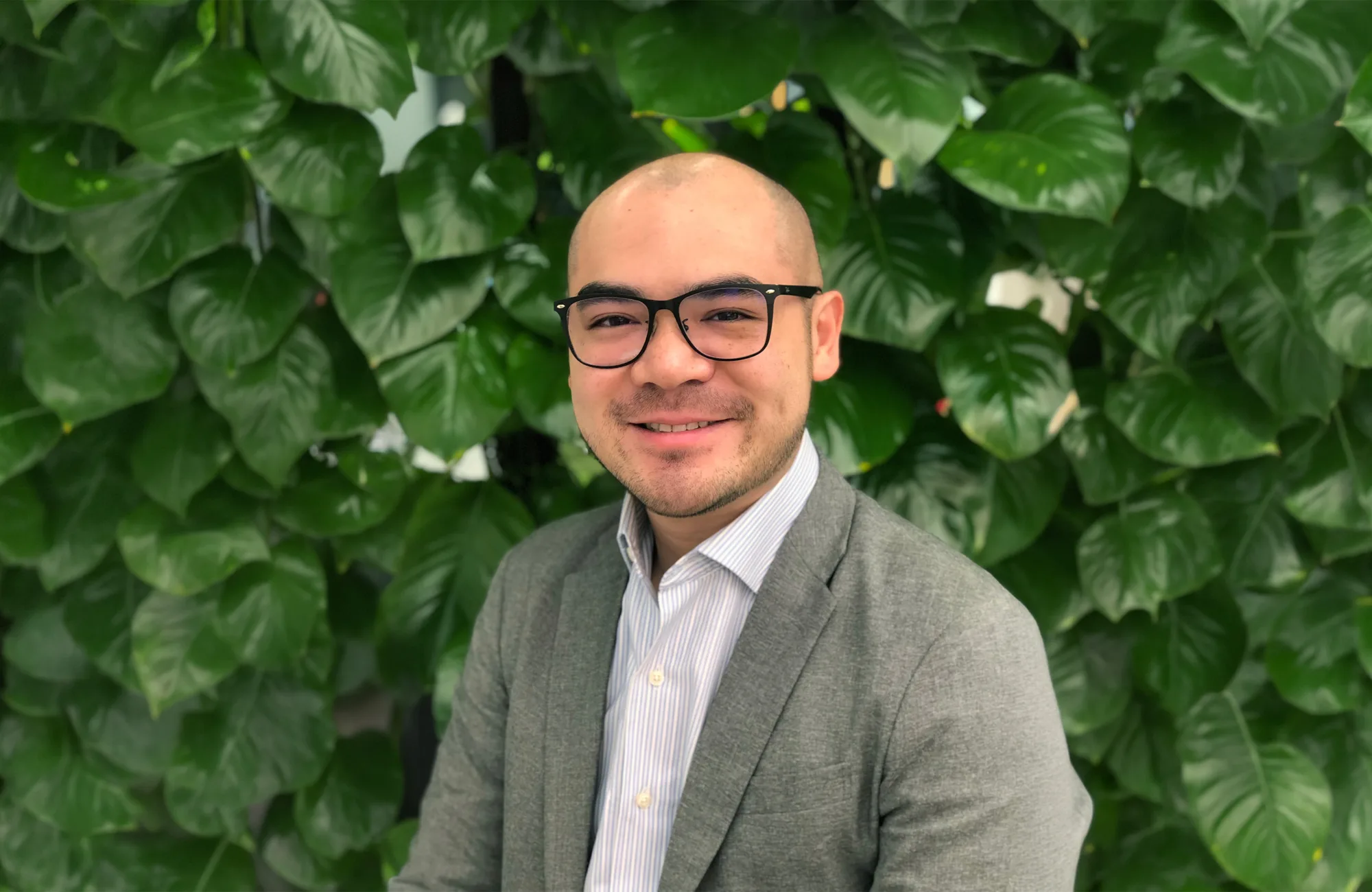 Portrait-style photo of a person wearing a light grey blazer over a white collared shirt, positioned in front of a dense green leafy wall. Upper body visible, with foliage filling the background.