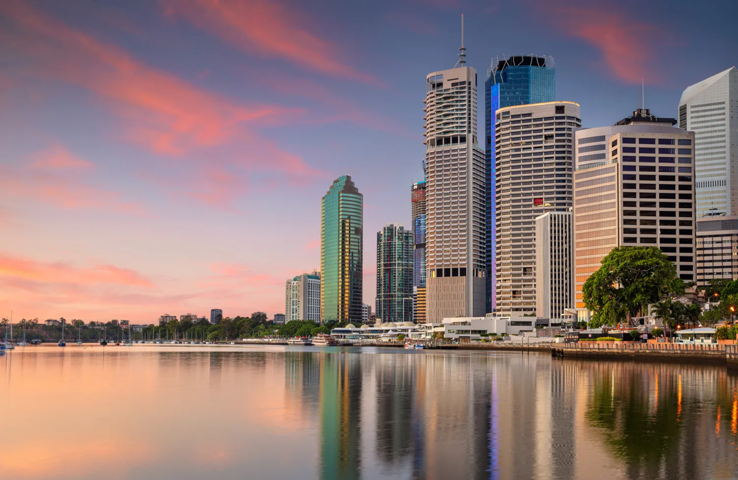 Tall modern skyscrapers with glass and concrete facades line a waterfront, reflecting in calm water under a pink and orange sunset sky. Trees and smaller buildings are visible near the shore.