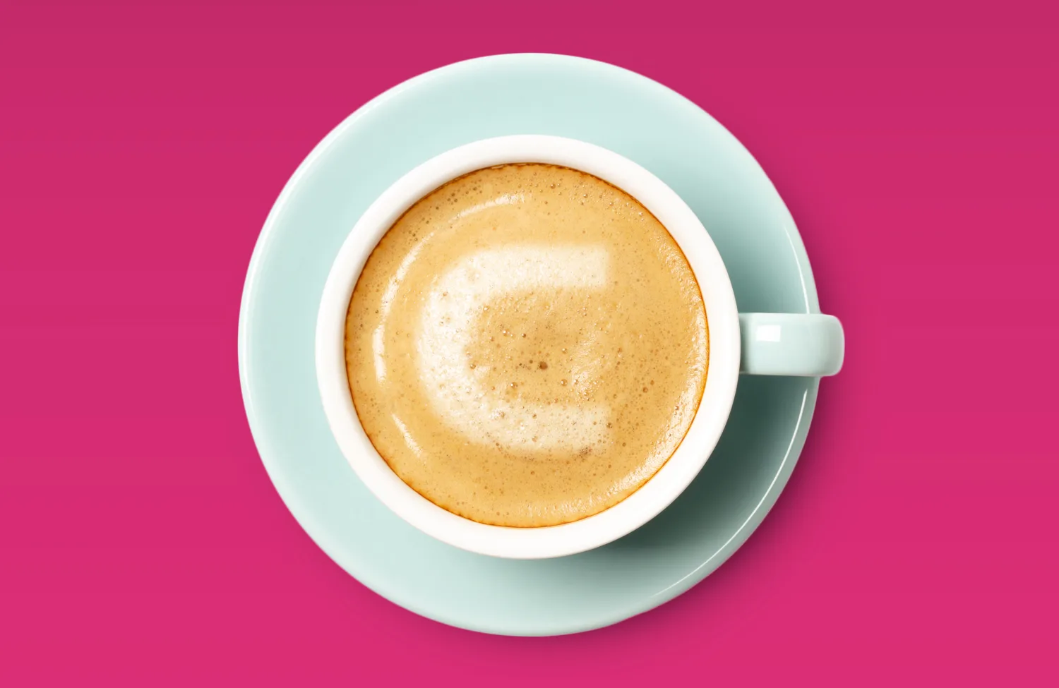 A top‑down view of a cup of frothy coffee in a light-blue saucer placed against a bright pink background. The foam on the coffee forms a C shape.