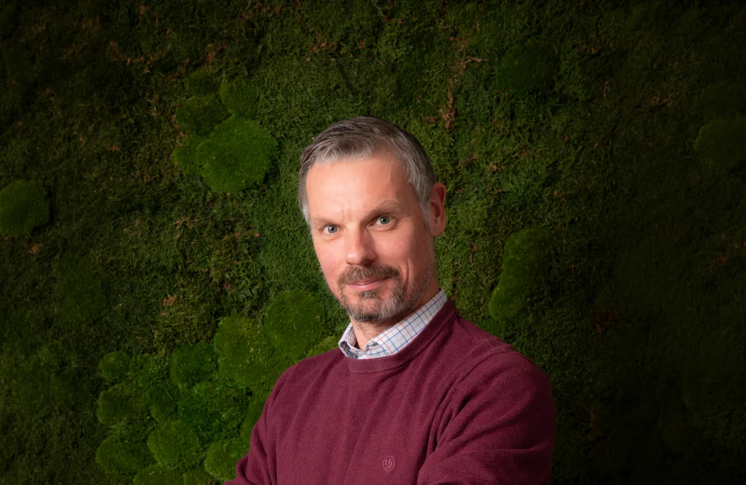 A person wearing a burgundy sweater over a checked shirt stands with folded arms in front of a textured green moss wall.