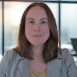 Upper‑body view of a person seated indoors, wearing a light grey jacket over a yellow top. The background shows a modern office setting with large windows, neutral walls, and soft daylight.