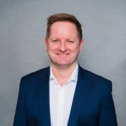 Studio head‑and‑torso portrait of a person standing against a plain grey textured backdrop, wearing a navy blue suit jacket over a white collared shirt, with hands loosely positioned in front.