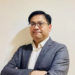 A person wearing glasses, a white buttoned shirt, and a gray blazer stands with arms crossed against a plain off‑white wall, framed from the waist up under indoor lighting.