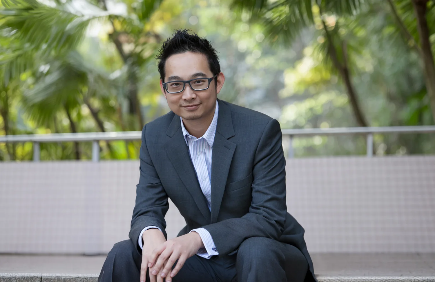A person in a dark grey business suit sits on a low ledge outdoors, hands clasped, with palm trees, greenery, and a tiled wall and railing in the background.