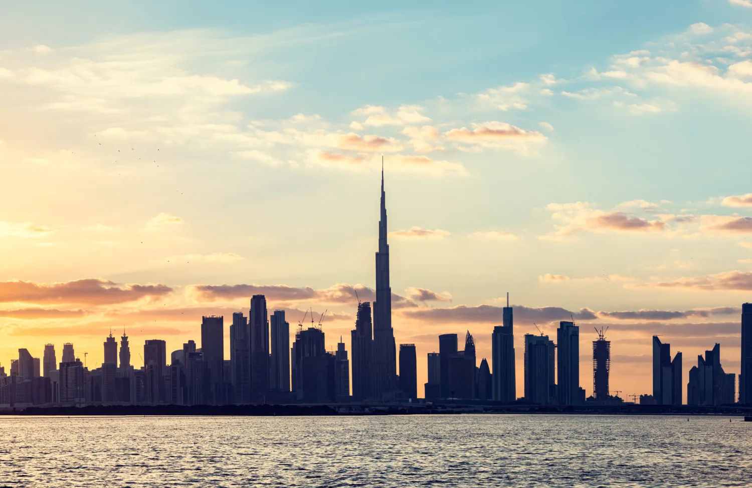 Dubai coastal skyline at sunset with numerous high‑rise buildings and a tall central tower silhouetted against the sky.