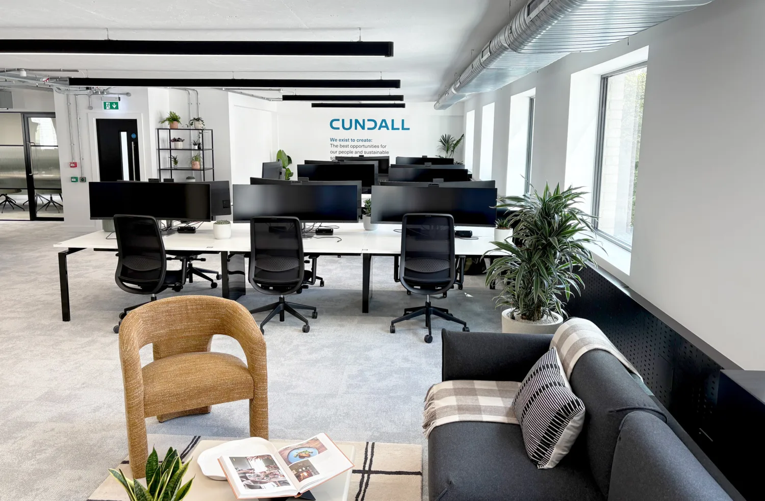 Modern office with workstations, black chairs, and large monitors. Foreground includes a brown chair, grey sofa with pillows and blanket, and open magazine. Wall text shows the Cundall logo and sustainability message. Potted plants and large windows.