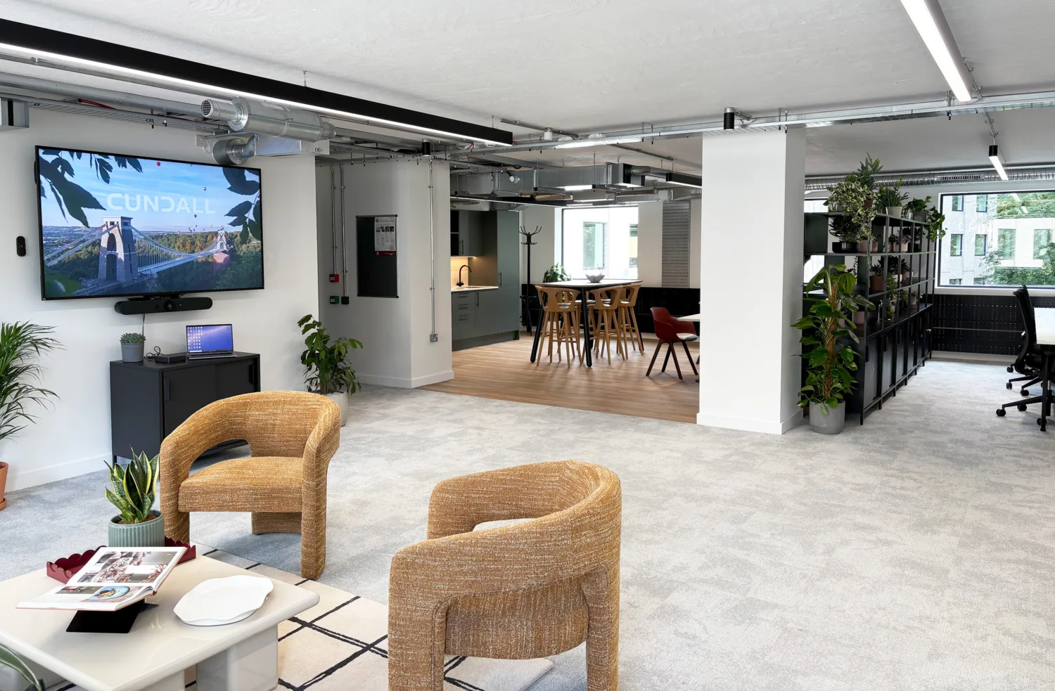 Modern office with a large screen showing “CUNDALL” and a bridge image. Includes wicker chairs, table with magazines, plants, laptop on cabinet, and open kitchen with bar stools.