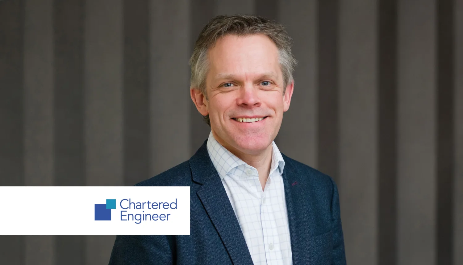 A person wearing a dark blue blazer over a white checkered shirt stands in front of a gray vertical-striped background. In the lower left corner, a white rectangle displays the logo and text “Chartered Engineer” with blue and green square icons.