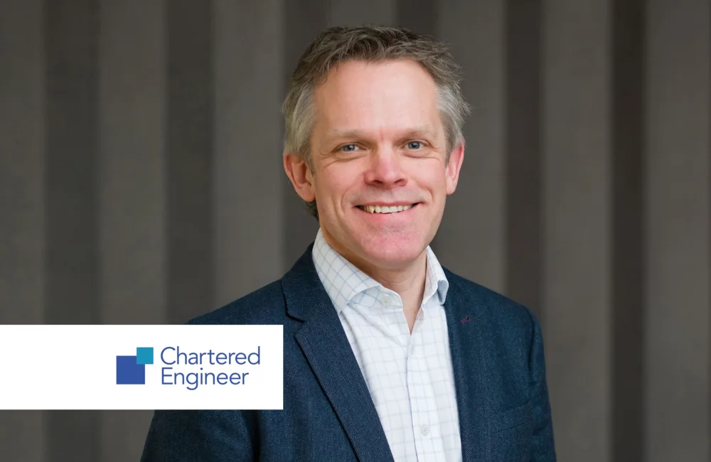 A person wearing a dark blue blazer over a white checkered shirt stands in front of a gray vertical-striped background. In the lower left corner, a white rectangle displays the logo and text “Chartered Engineer” with blue and green square icons.