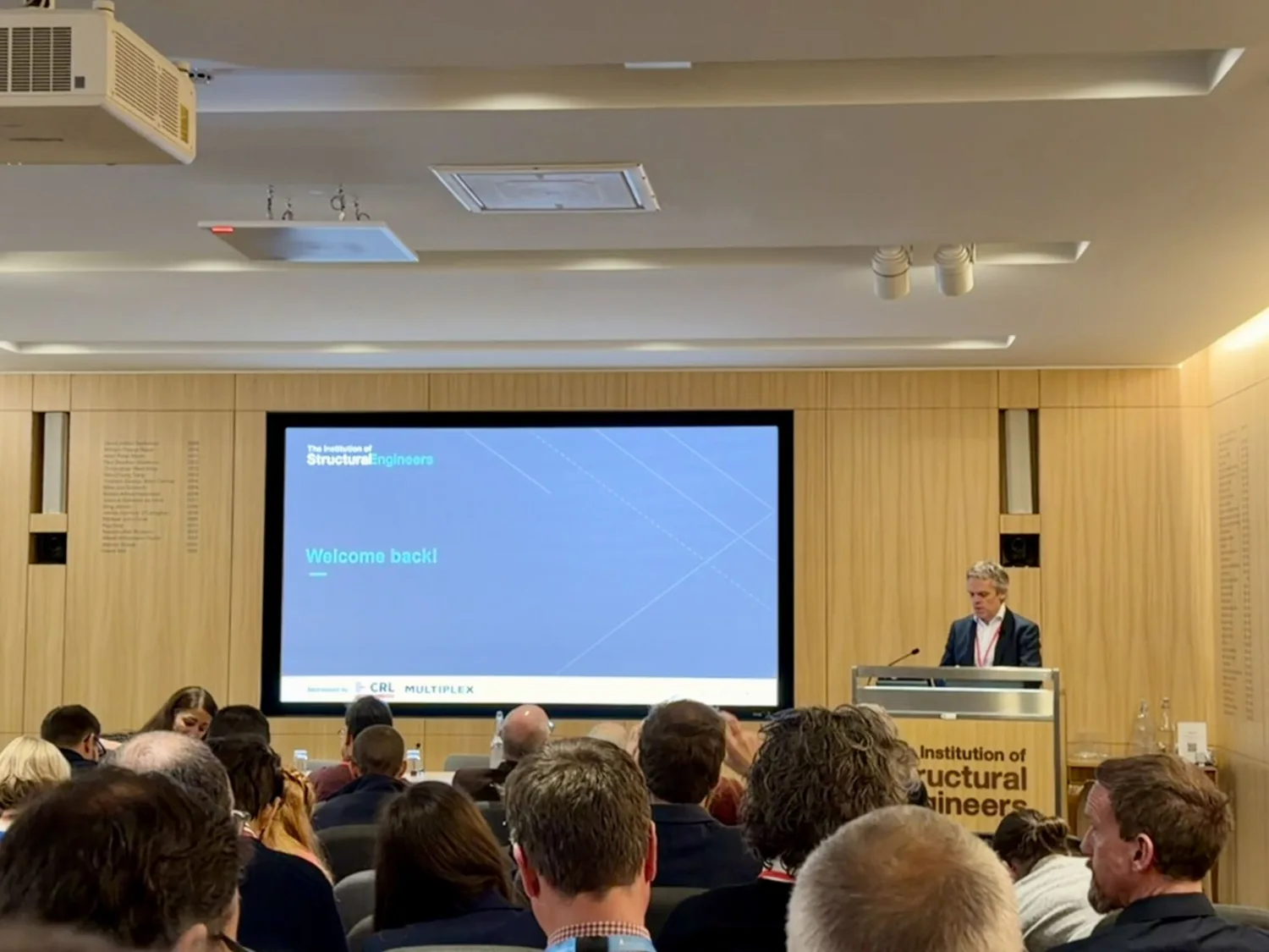 A conference room with attendees seated facing a large screen displaying the text ‘Welcome back!’ and the logo of the Institution of Structural Engineers. A person stands at a podium labelled with the same institution name.